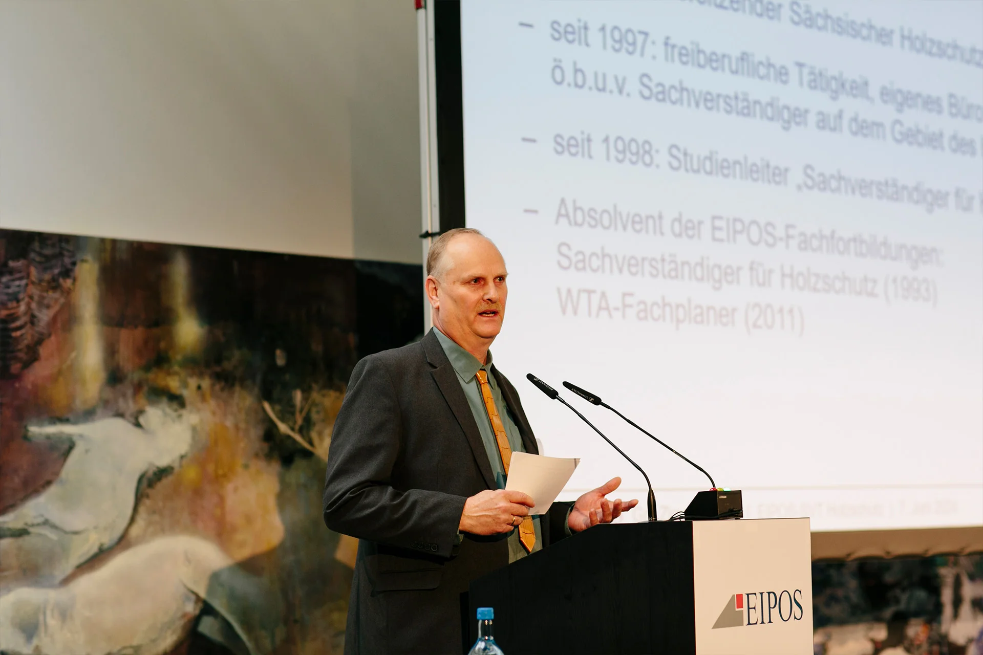 Aufnahme eines älteren Refernten am Rednerpult mit der Vorstellungsfolie im Hintergrund auf der Leinwand