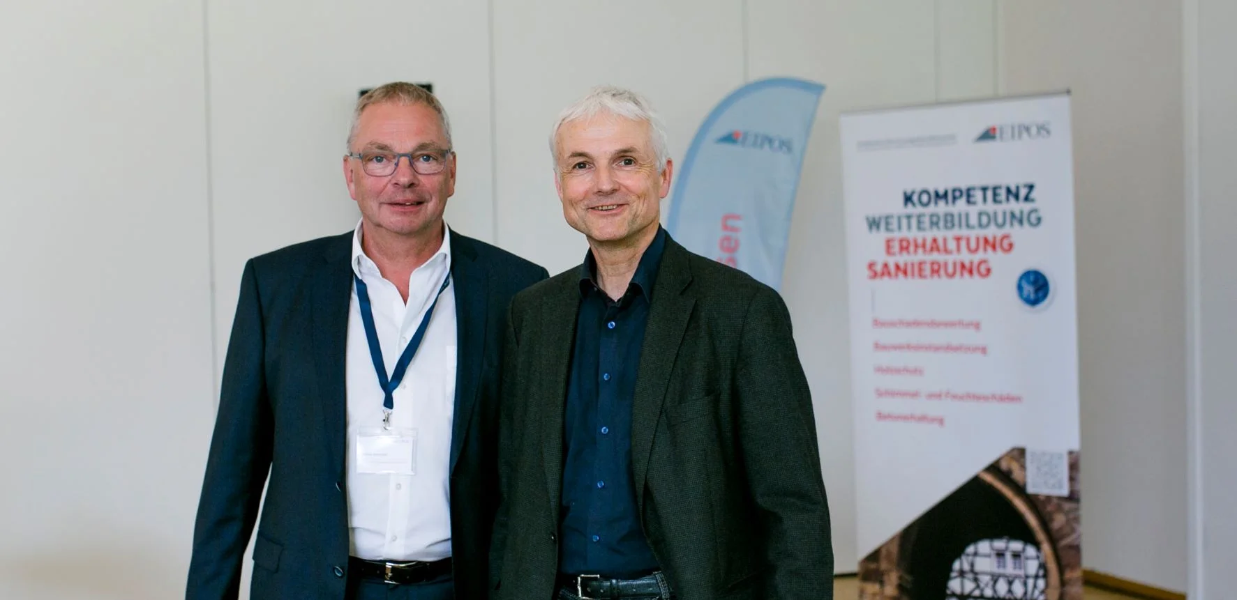 Zwei Herren im Anzug an der Bühne. Im Hintergrund ist ein Aufsteller zu Weiterbildungen Erhalten und Sanieren