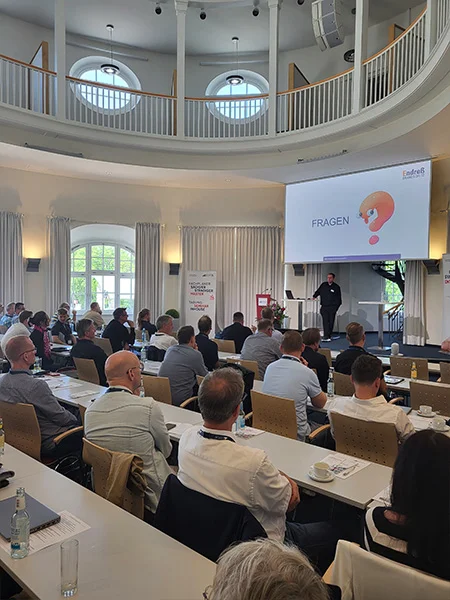 Leicht schräger Blick über die Zuhörenden zur Bühne mit Leinwand